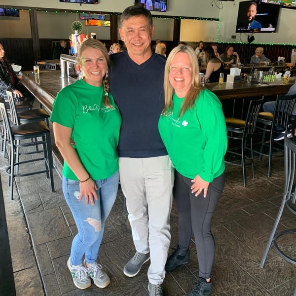 Two women in green shirts and a man in a blue sweater smiling inside The Tavern at Sugar Hill.