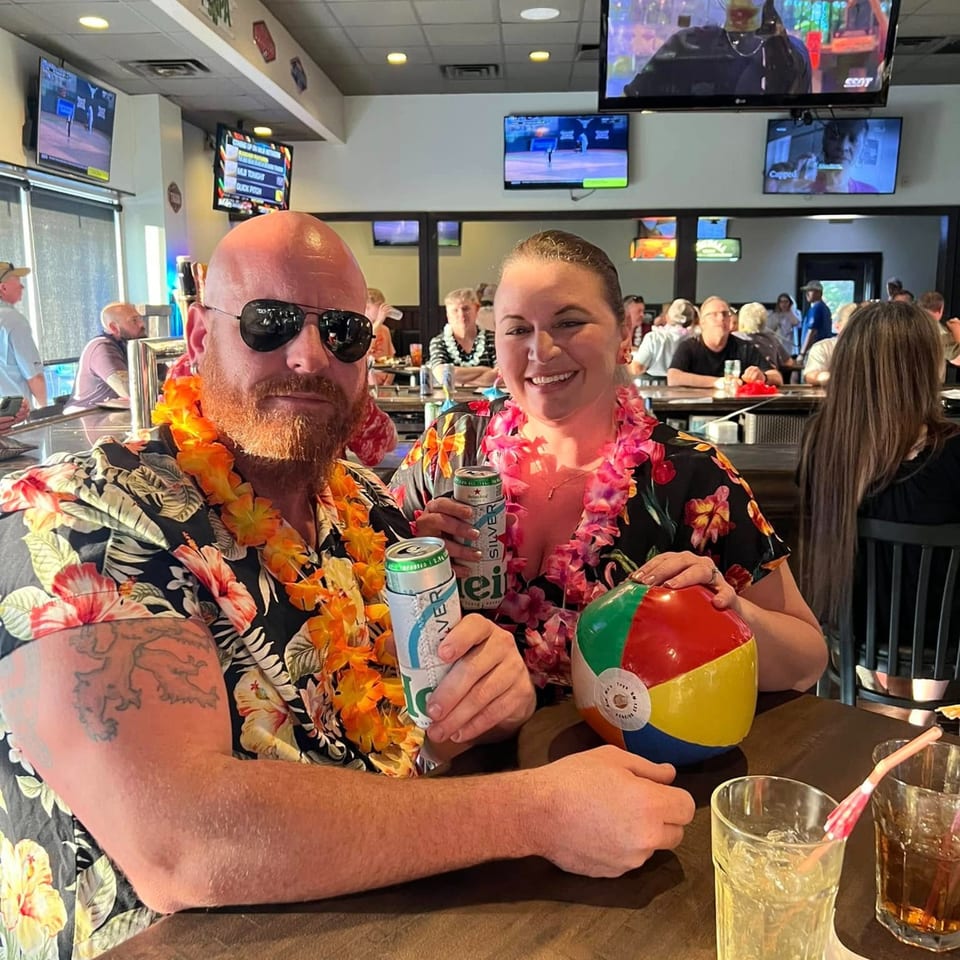 Two patrons in Hawaiian shirts and leis smile at the bar, holding drinks and a beach ball.